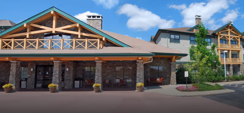 Front entrance of a lodge-style senior living building with stone pillars, covered porch seating, planters, and a landscaped driveway under a blue sky.