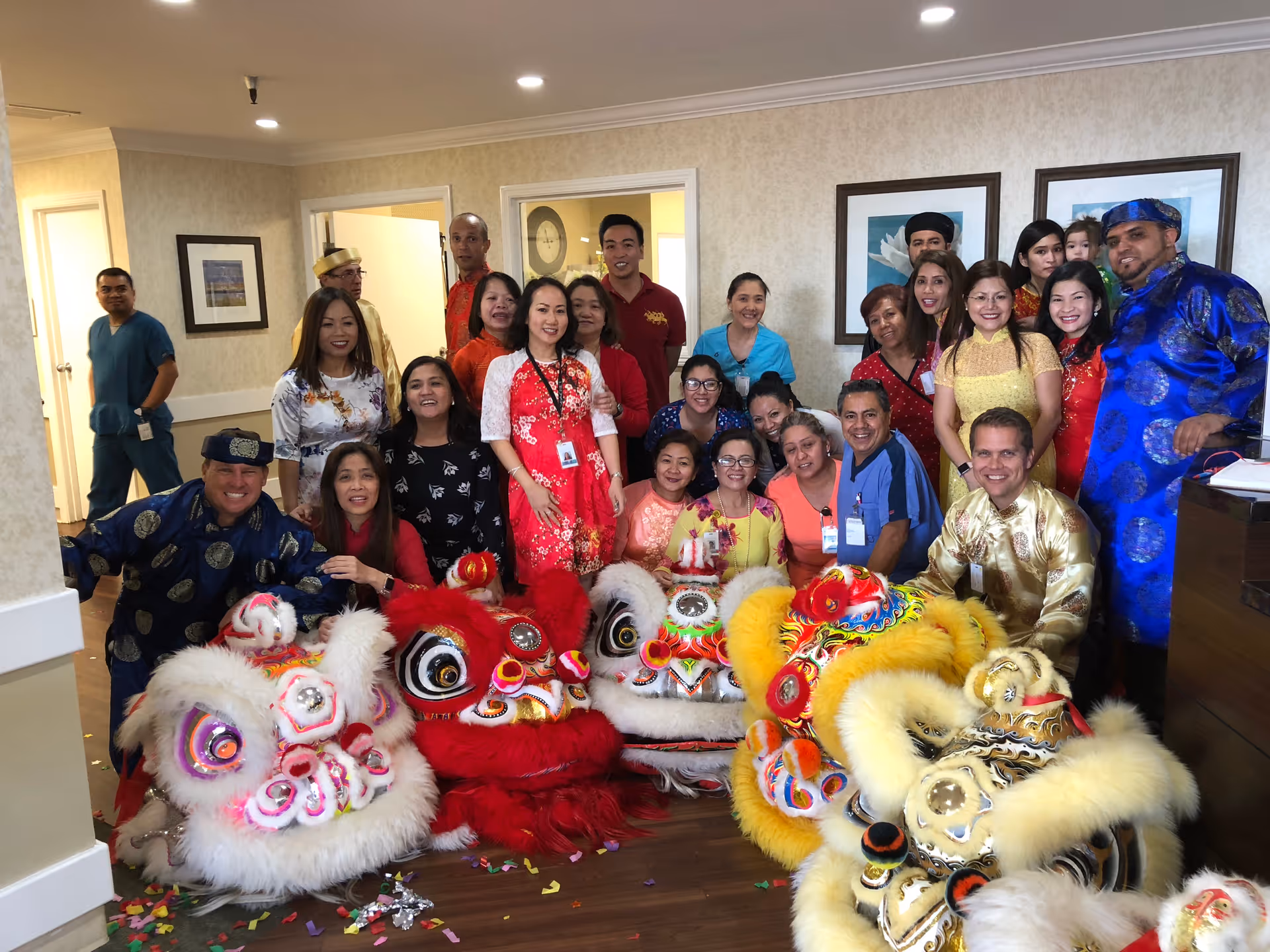 A group of people posing indoors at Mission Palms Healthcare Center, dressed in colorful traditional clothing, with four vibrant lion dance costumes in front of them on the floor. The setting appears to be a common area or lounge with framed artwork on the walls and a wooden floor.