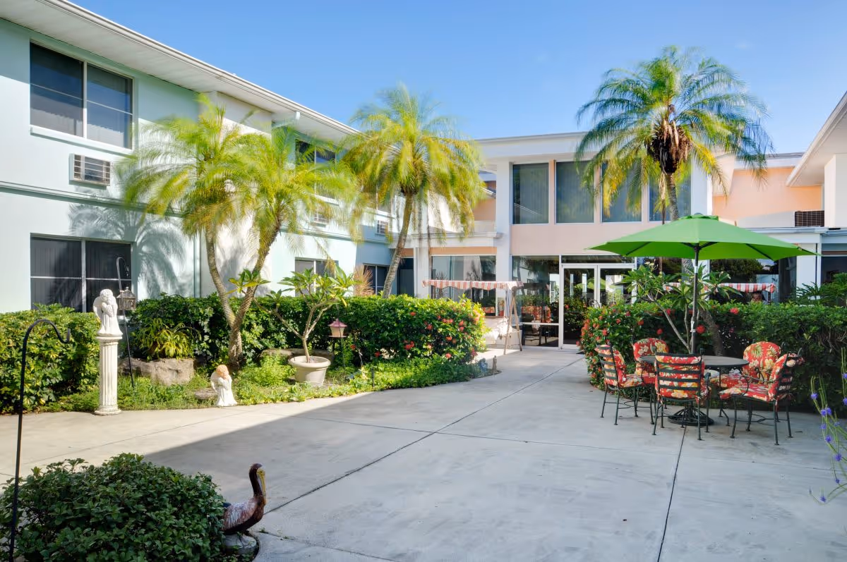 Sunny landscaped courtyard of a senior living facility with palm trees, a paved walkway and patio tables and chairs under a green umbrella in front of the building entrance.