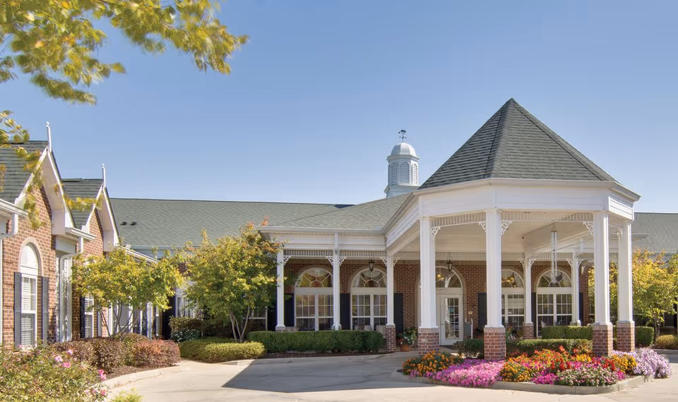 Brick senior living facility with a white-columned front porch and vibrant flower beds.