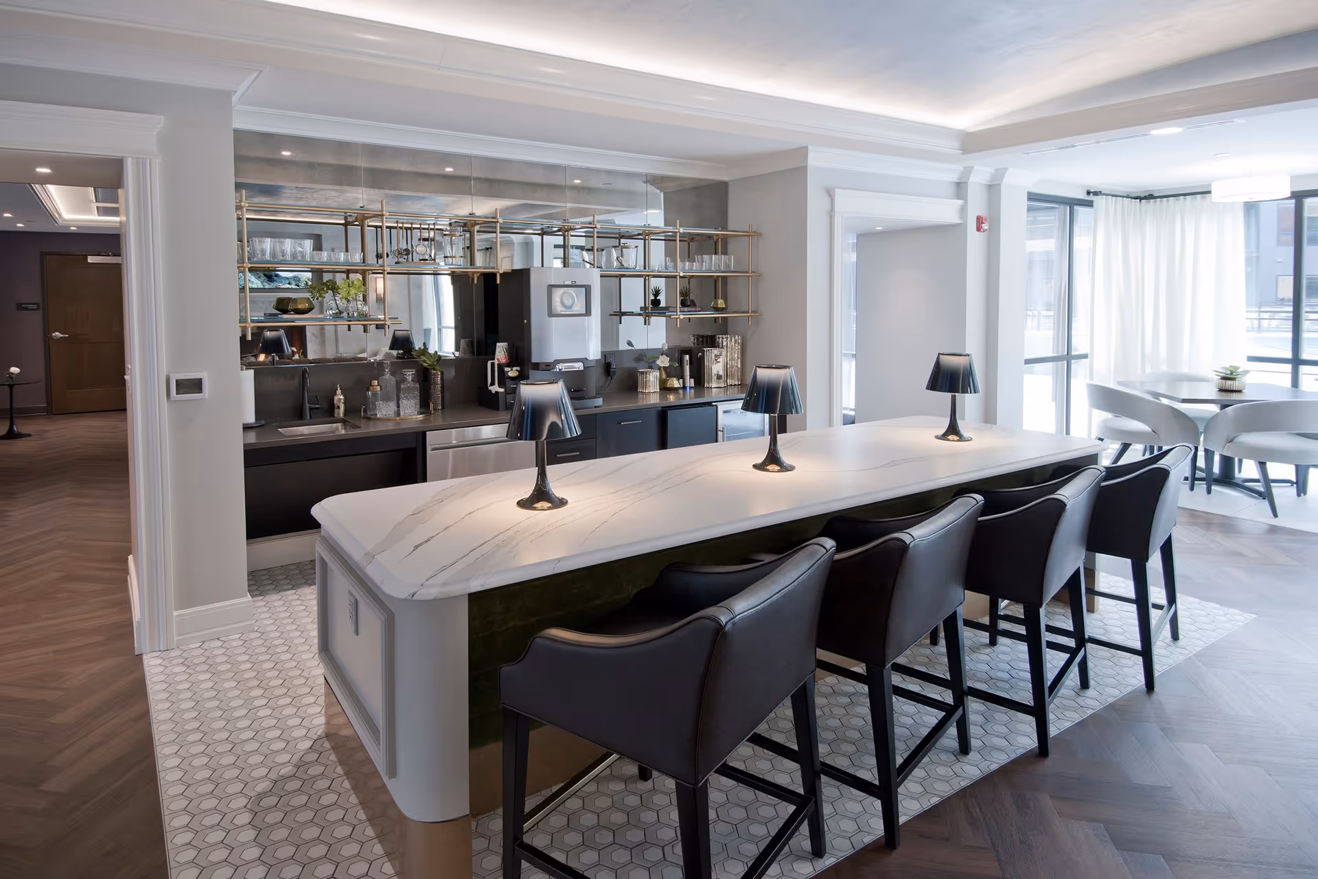 Modern kitchen area with a long white marble island countertop featuring four black chairs and three small black lamps. Behind the island is a sleek kitchen setup with a mirrored backsplash, open shelving with glassware, and various kitchen appliances. To the right, there is a dining area with a round table and chairs near large windows with white curtains allowing natural light to fill the space.