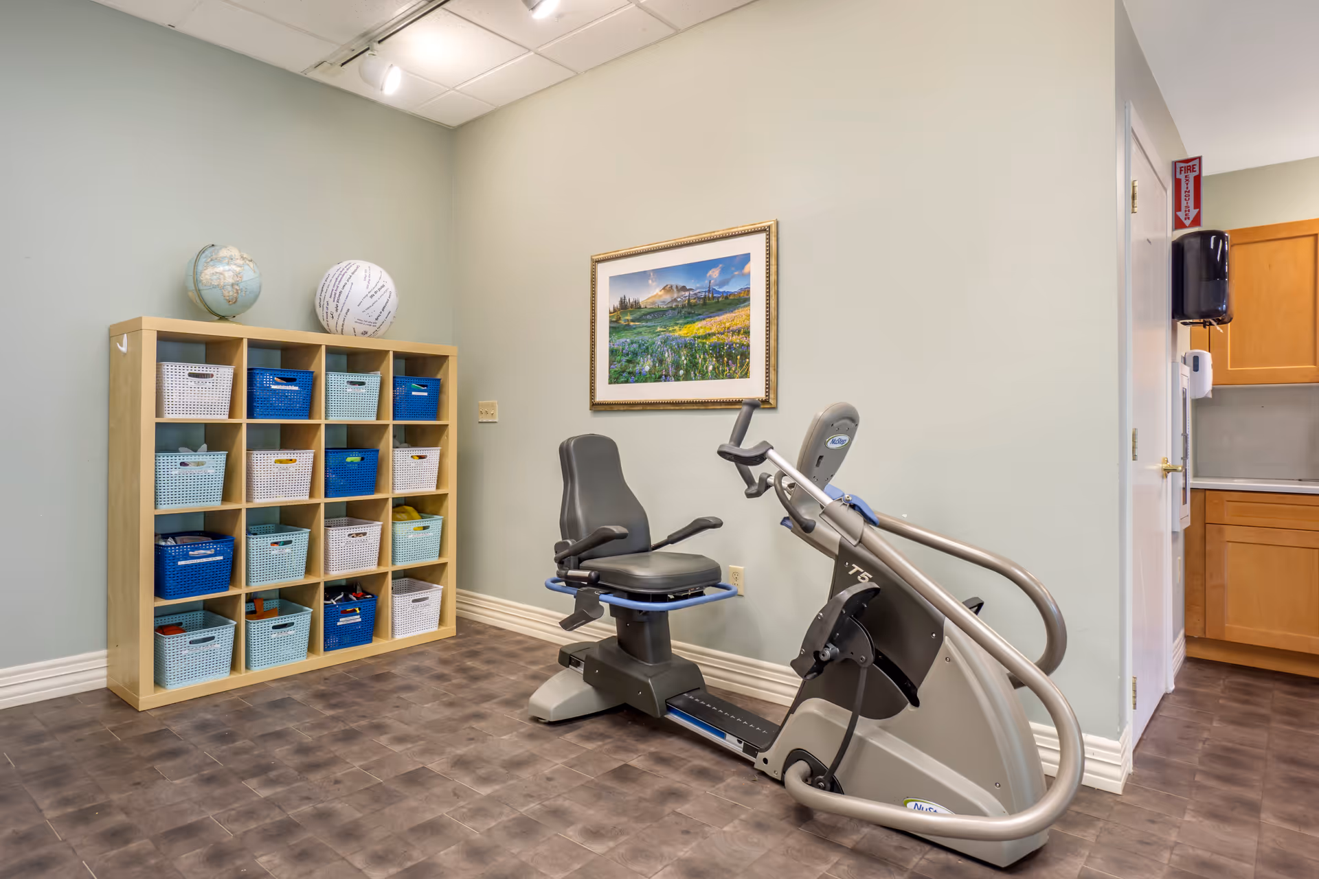 A small exercise room with a recumbent exercise bike positioned near a wall. Next to the bike is a wooden cubby shelf filled with blue, white, and light blue storage baskets. On top of the shelf are a globe and a white ball with text. A framed landscape photograph hangs on the wall above the exercise bike. The room has light green walls and a brown floor, with a partial view of a kitchen area to the right.