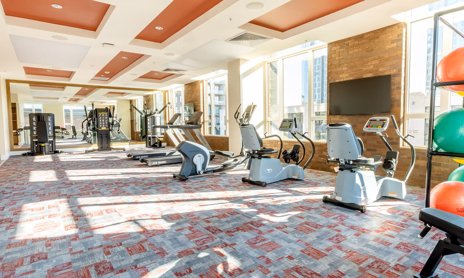 Bright and spacious fitness room with large windows allowing natural light to fill the space. The room features exercise equipment including treadmills, stationary bikes, weight machines, and a rack with colorful exercise balls. The ceiling has recessed lighting with a modern design, and the floor is covered with a patterned carpet in shades of red and gray.
