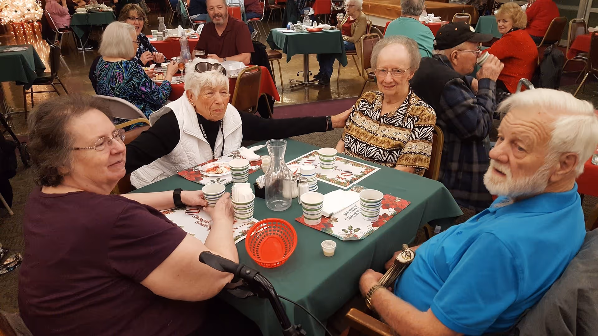 Several elderly residents sit around a holiday-decorated dining table with cups, plates and a pitcher at a communal meal.