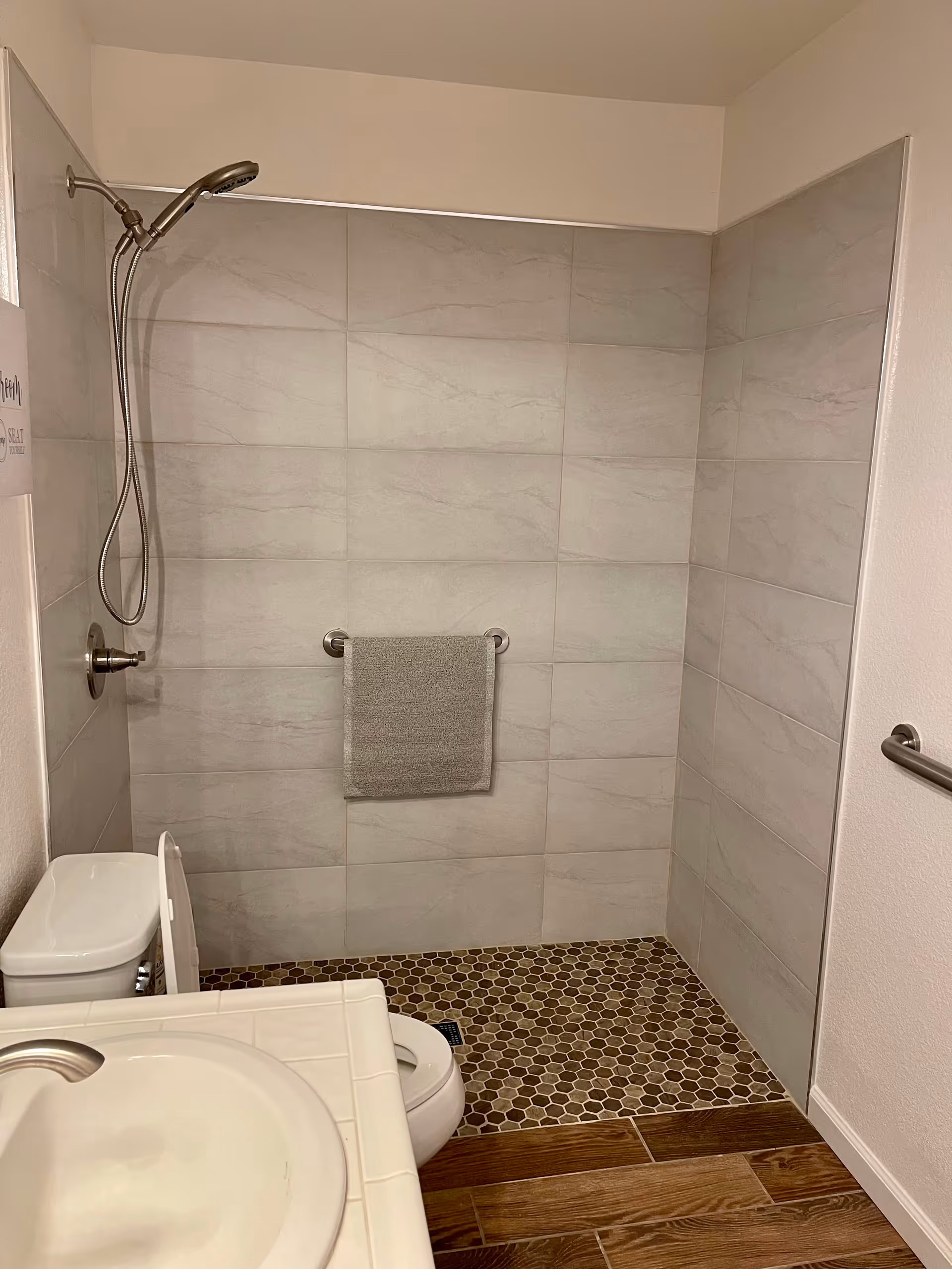 A bathroom with a walk-in shower featuring large light gray tiles on the walls and small hexagonal brown tiles on the floor. There is a silver showerhead with a flexible hose, a towel rack with a gray towel, a white toilet, and a white sink with a silver faucet. The floor outside the shower is wood-patterned tile.