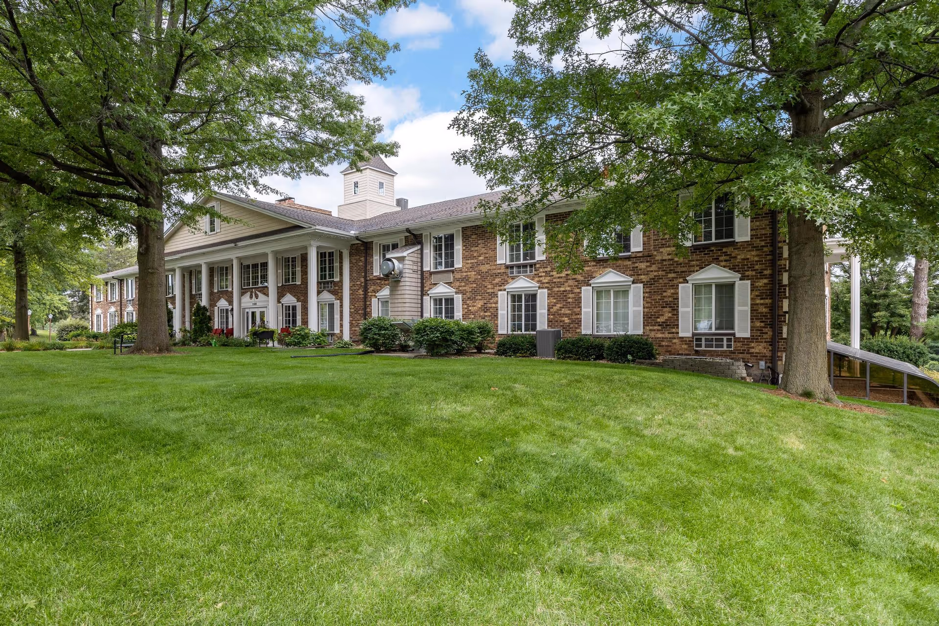 Brick two-story senior living building with white columns, many windows, and a large grassy lawn framed by trees.