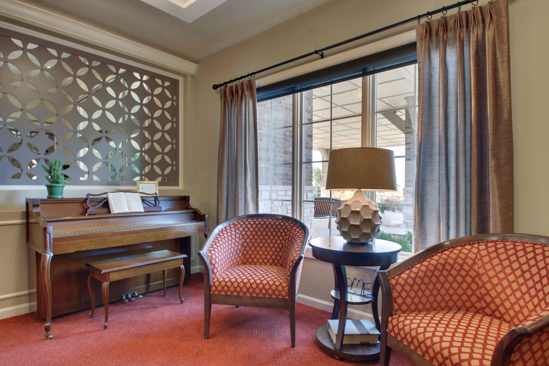 A cozy sitting area in a senior living facility featuring a wooden upright piano with an open book on the music stand, two patterned armchairs with red and beige upholstery, a round side table with a decorative lamp, and a large window with beige curtains overlooking an outdoor patio.