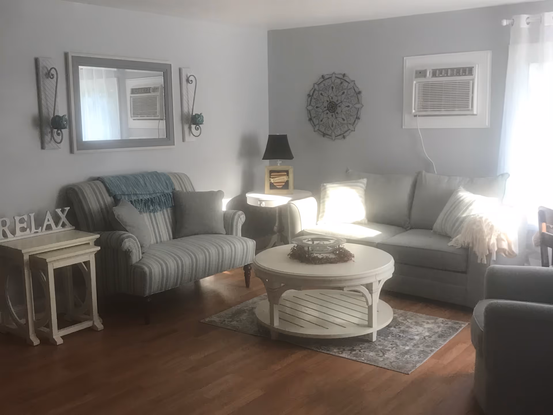 A cozy living room with a gray couch, a striped loveseat, a round white coffee table on a patterned rug, a side table with a lamp, wall decorations including a mirror and a decorative metal piece, and an air conditioning unit in the window.