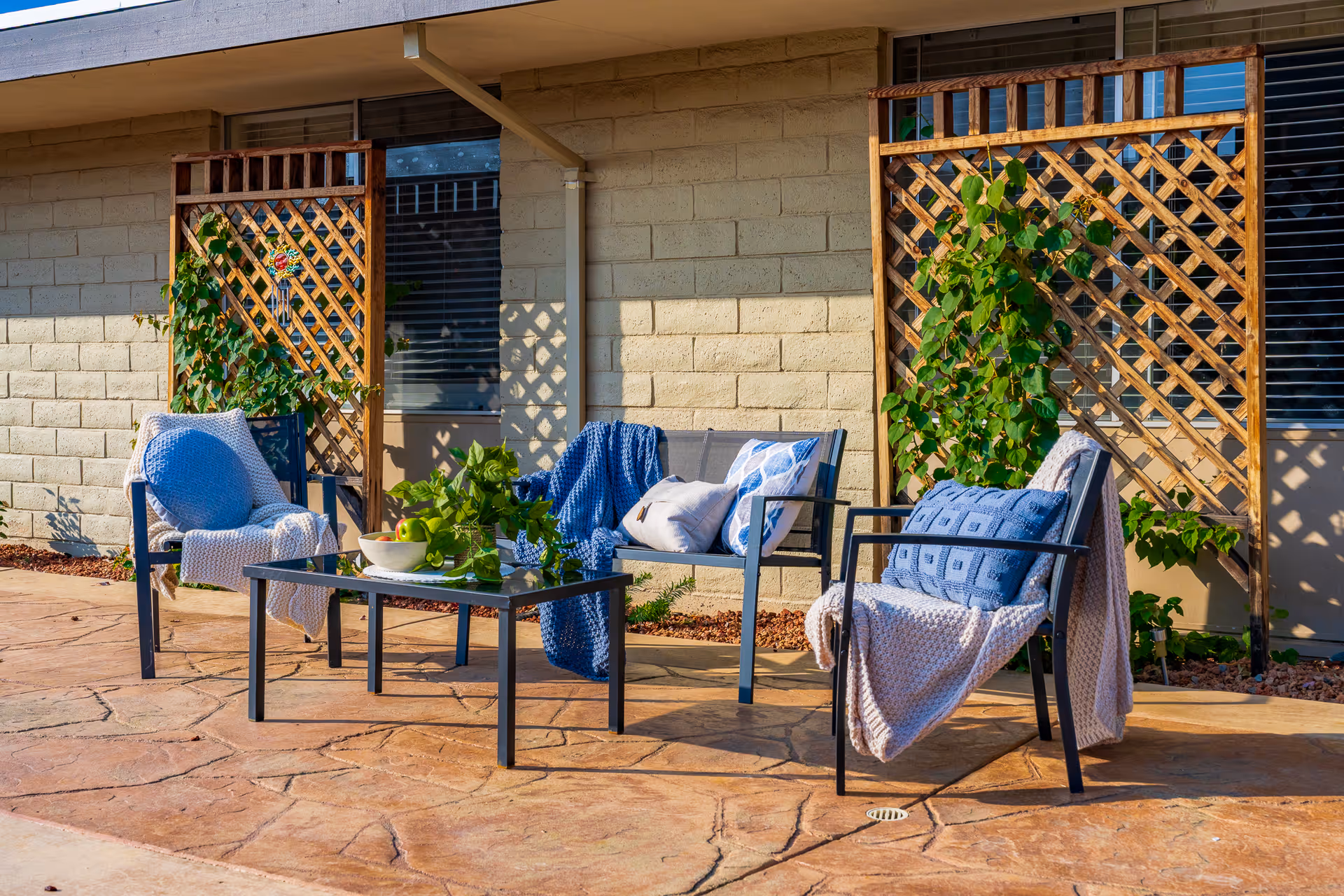 Sunlit outdoor patio with three cushioned chairs and a coffee table in front of a building wall with trellis and climbing plants.