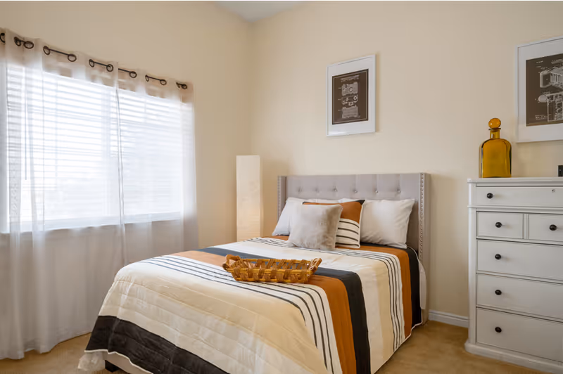 A cozy bedroom with a neatly made bed featuring a striped comforter in shades of white, orange, and black. The bed has a tufted headboard and several pillows. A wicker tray is placed on the bed. To the right, there is a white dresser with multiple drawers and a large amber glass bottle on top. Two framed artworks hang above the bed and dresser. A tall floor lamp stands in the corner near a window with sheer white curtains allowing natural light to fill the room.