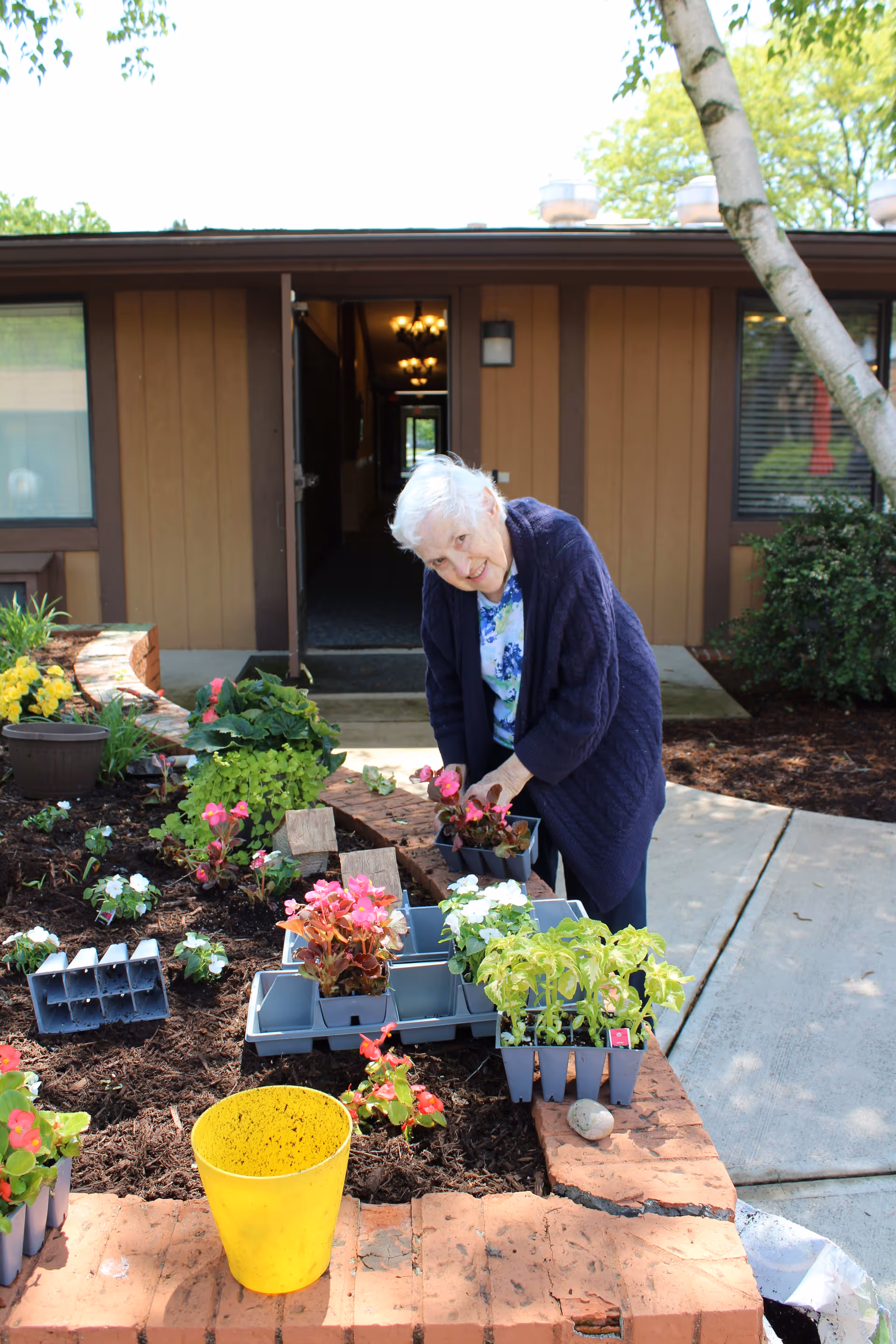 An elderly woman with white hair wearing a navy blue cardigan and floral shirt is gardening in a raised brick flower bed outside a building. She is planting pink and white flowers, with several trays of plants and a yellow bucket nearby. The building behind her has brown siding and an open door leading to a hallway with chandeliers.