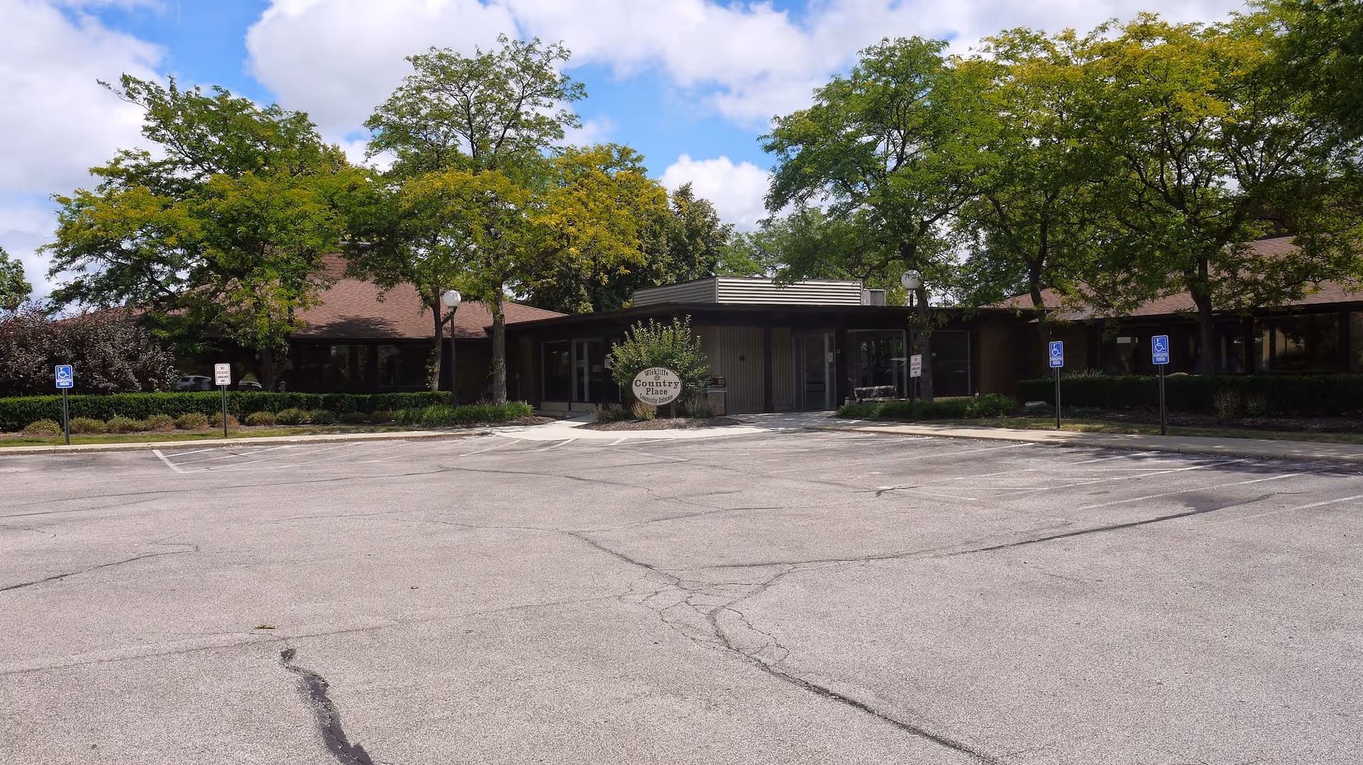 Exterior view of Wickliffe Country Place facility with a large empty parking lot in front, surrounded by trees and greenery under a partly cloudy sky.