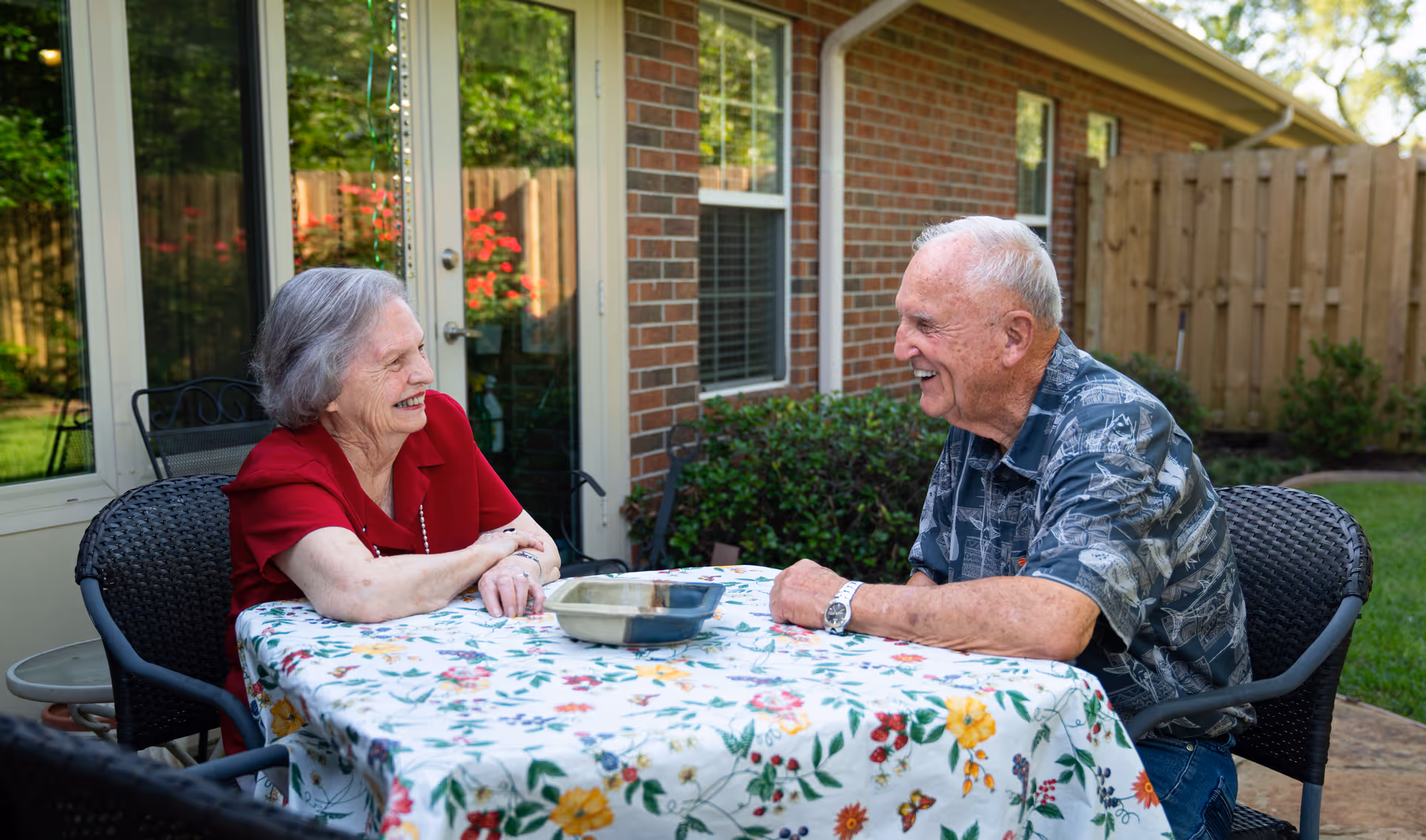 An elderly woman and an elderly man sitting at a table covered with a floral tablecloth outdoors, smiling and engaging in conversation. They are seated on black wicker chairs near a brick building with windows and a wooden fence in the background.