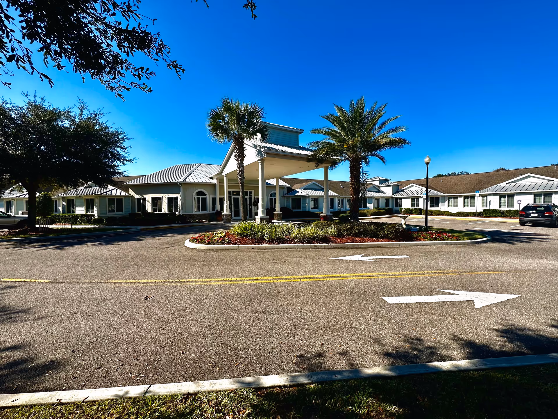 Front exterior view of Lady Lake Senior Living facility with a covered entrance, palm trees, landscaped flower beds, and a clear blue sky.