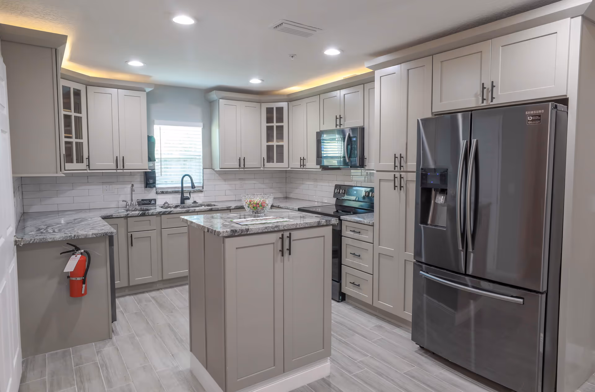 Modern kitchen with gray cabinets, a central island with marble countertop, stainless steel refrigerator, stove, microwave, and a window above the sink. The kitchen has tiled flooring and recessed lighting with under-cabinet lighting.