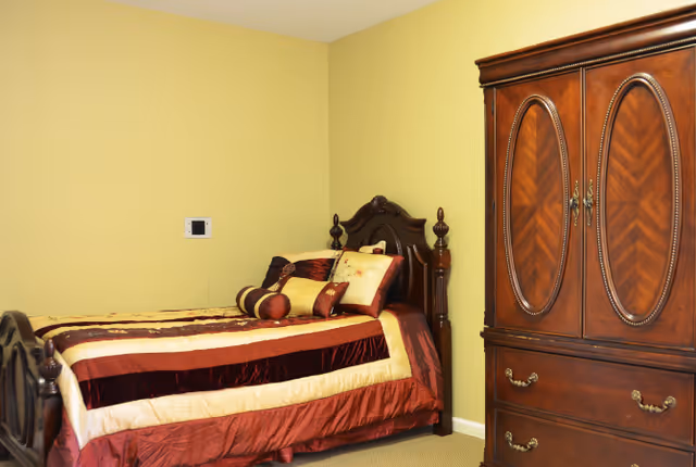 A bedroom with a single wooden bed featuring an ornate headboard and footboard, dressed with a striped bedspread in shades of red, gold, and cream, along with several decorative pillows. Next to the bed is a large wooden armoire with detailed carvings and brass handles. The walls are painted yellow and the floor is carpeted.