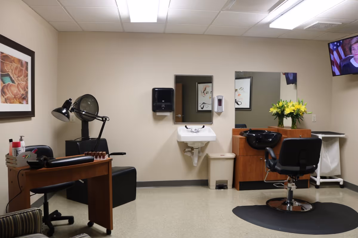 Interior of a senior housing salon room with a black salon chair in front of a wooden counter with a black wash basin. There is a sink with a mirror above it on the wall, a paper towel dispenser, and a soap dispenser. A small wooden desk with a black lamp, nail polish bottles, and other salon supplies is on the left side. A TV is mounted on the upper right corner of the wall, and a vase with yellow flowers is on the counter. The room has beige walls and a tiled floor.
