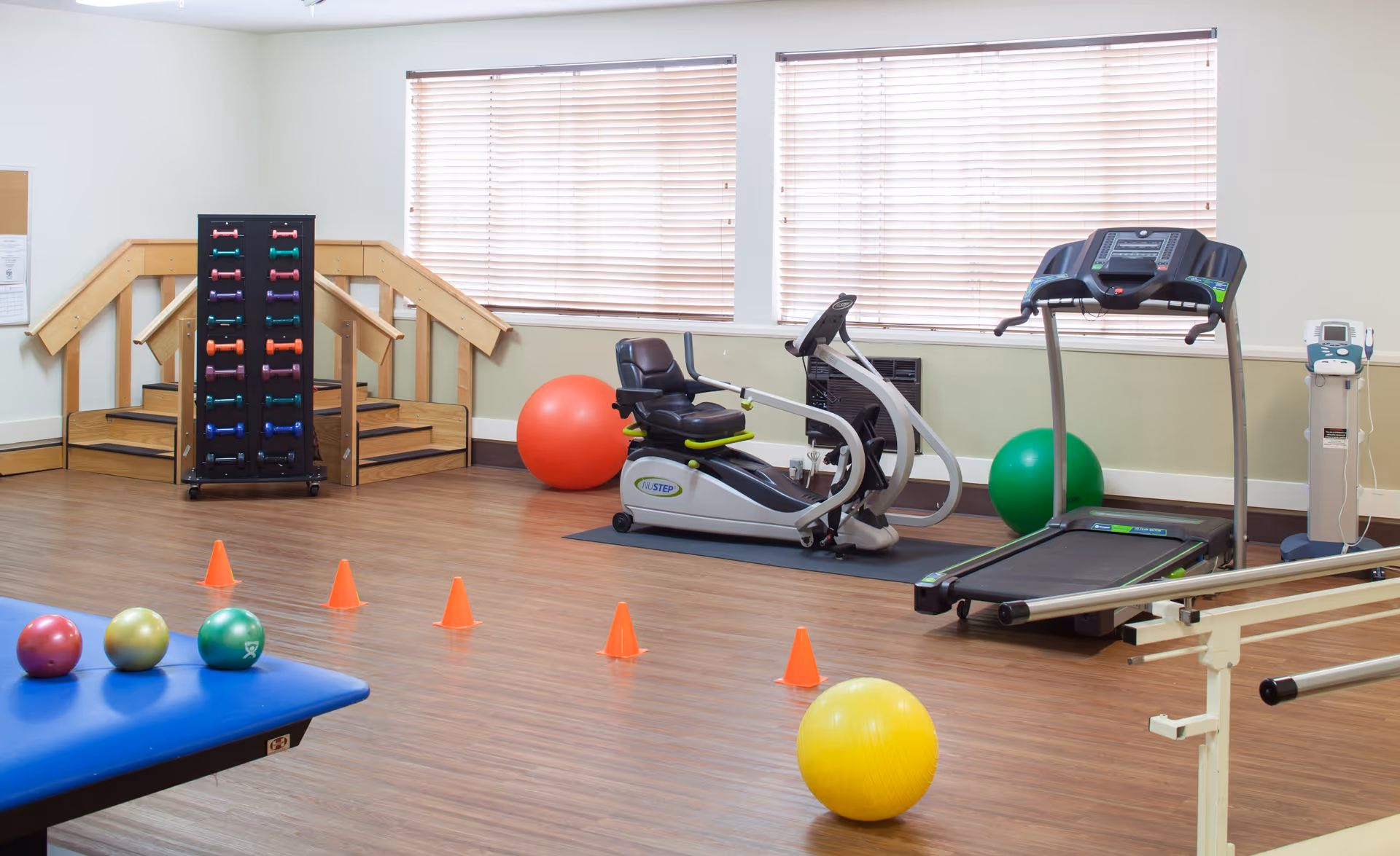 A fitness room with exercise equipment including a recumbent bike, treadmill, colorful dumbbells on a rack, exercise balls, orange cones, and parallel bars. The room has wooden flooring and windows with blinds.
