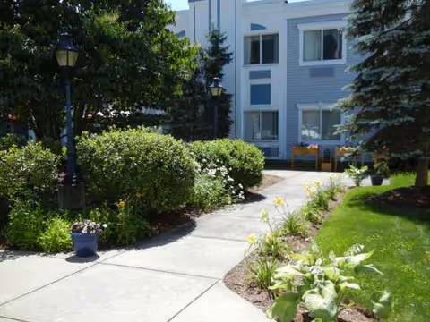Outdoor walkway with shrubs and flowers beside a light-blue senior living building.