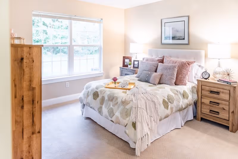 A bright and cozy bedroom with a large window letting in natural light. The bed is made with a patterned comforter and several decorative pillows in shades of pink and gray. There is a wooden nightstand on each side of the bed, each with a lamp and decorative items including a clock and picture frames. A wooden dresser is partially visible on the left side of the image. The walls are painted a soft beige color and a framed picture hangs above the bed.
