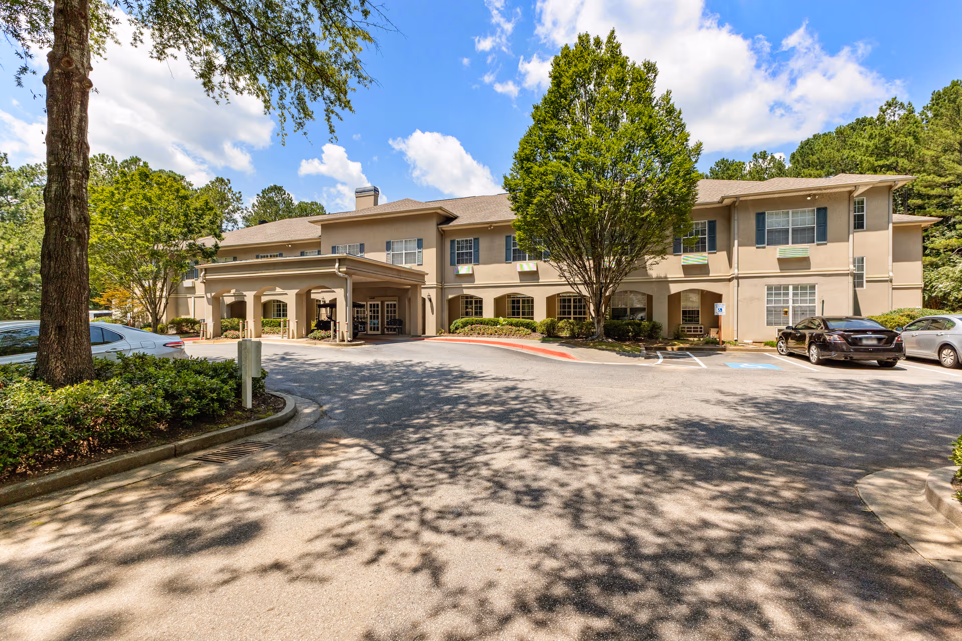 Exterior view of The Reserve at Peachtree City Assisted Living and Memory Care building with a covered entrance, several parked cars, and trees casting shadows on the driveway under a partly cloudy sky.