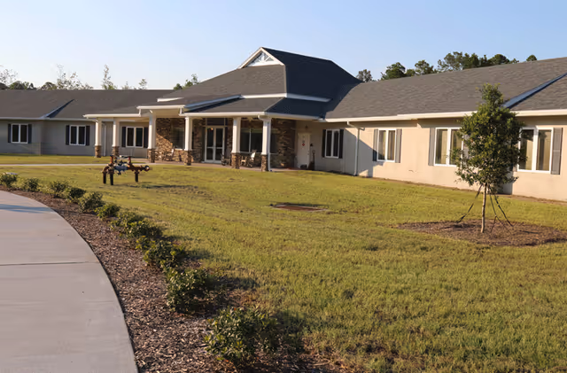 Exterior view of a single-story senior living facility building with a covered entrance, surrounded by a grassy lawn and small shrubs along a curved sidewalk under a clear sky.