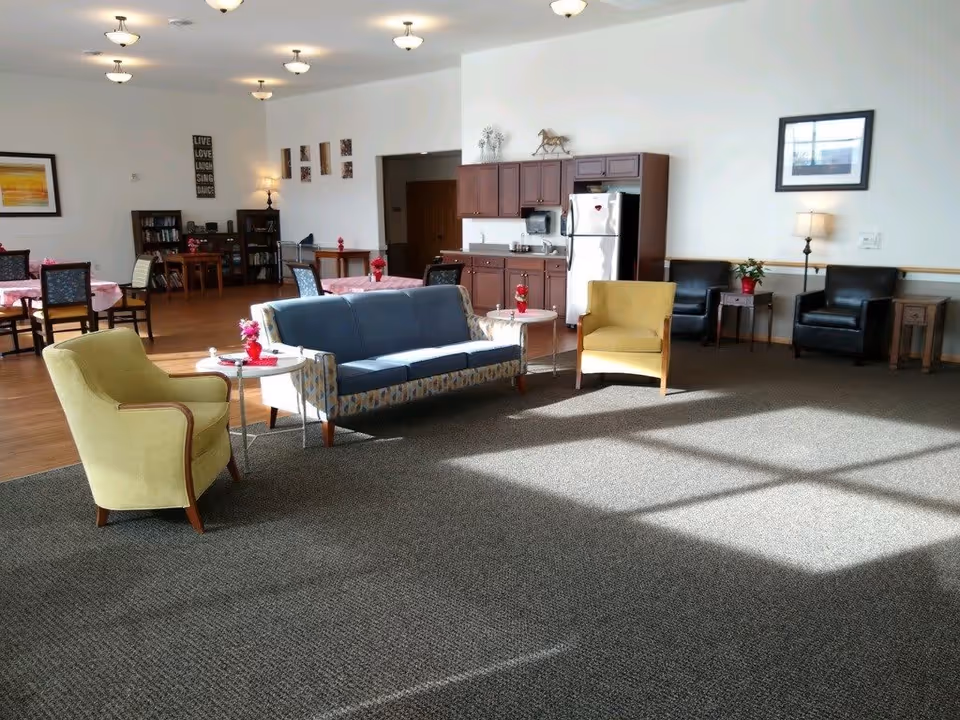 A spacious senior living common area with a mix of seating including a blue sofa, two yellow armchairs, and two black armchairs. There are small tables with red flower arrangements, a kitchenette with wooden cabinets and a stainless steel refrigerator, and dining tables with pink tablecloths in the background. The room is well-lit with ceiling lights and natural light from windows.