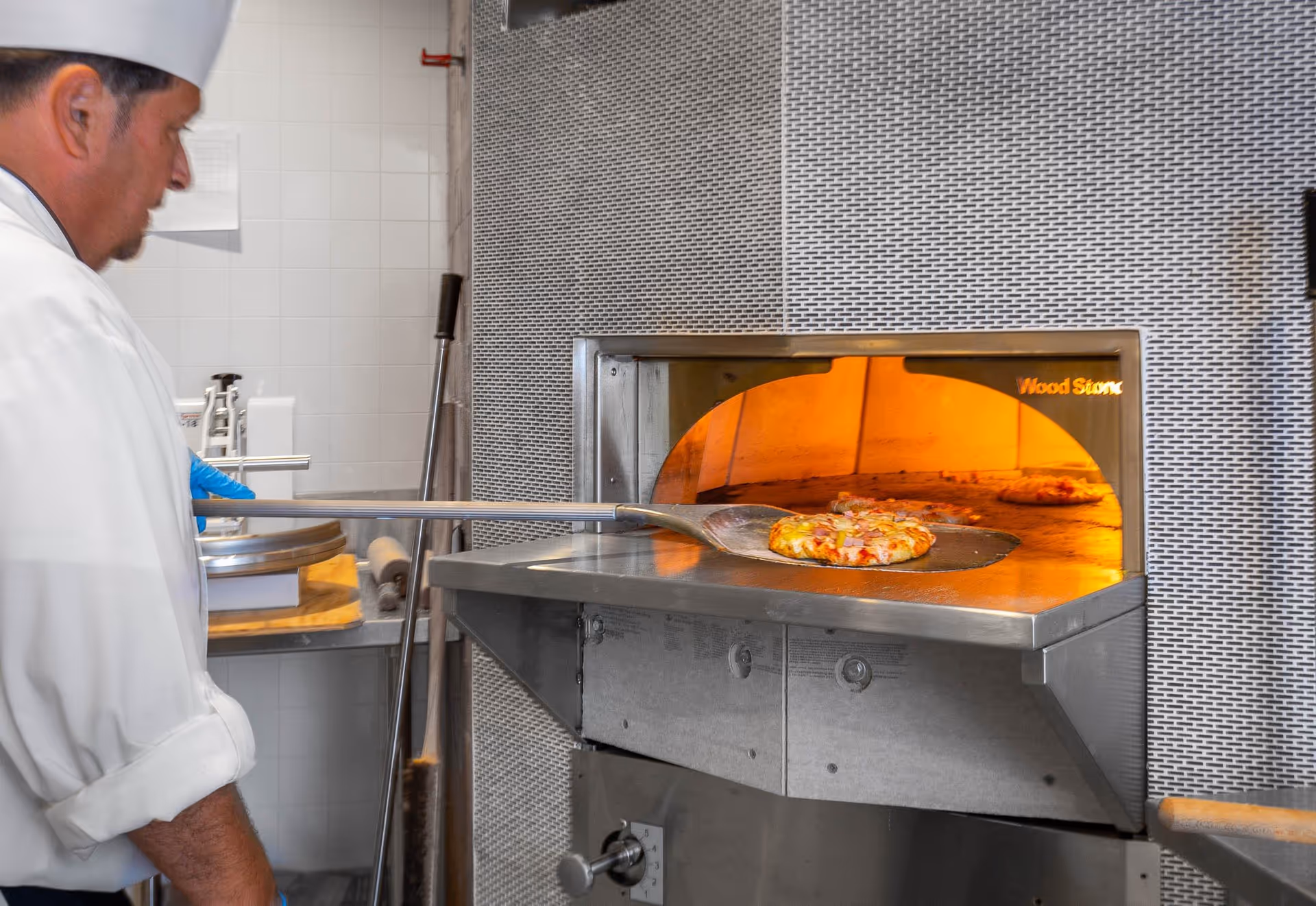 A chef wearing a white uniform and hat is using a pizza peel to place or remove a pizza from a commercial pizza oven with an orange glowing interior.