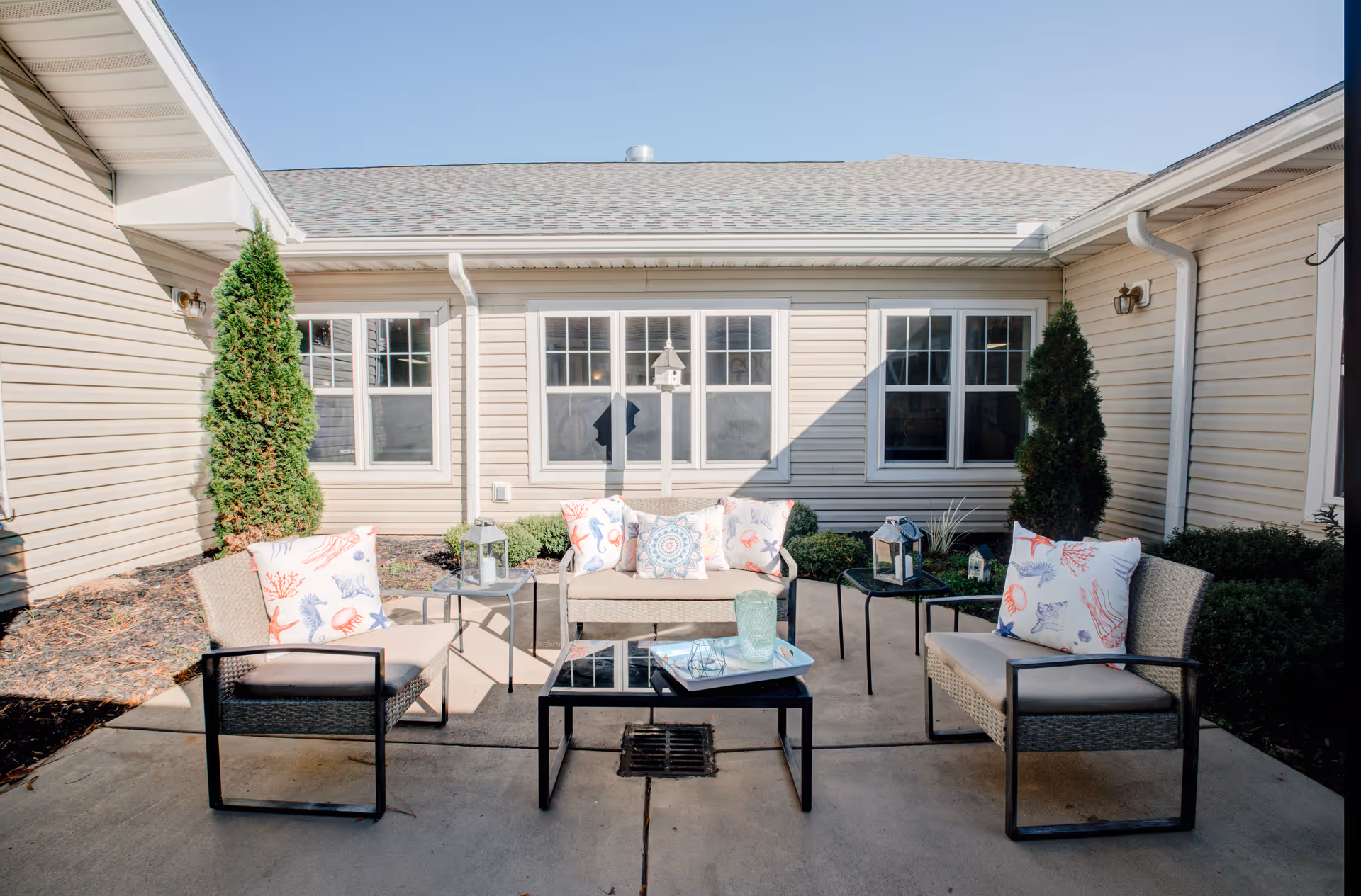 Outdoor patio area with beige cushioned wicker chairs and a loveseat arranged around a black glass coffee table. The seating has decorative pillows with sea-themed patterns. The patio is surrounded by beige siding walls with windows and small green shrubs and trees.