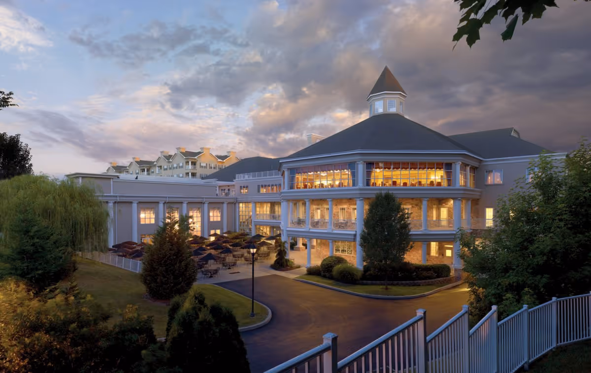 Evening exterior view of a large illuminated multi-story senior living building with a turret, wraparound balconies, patio umbrellas, and a driveway.