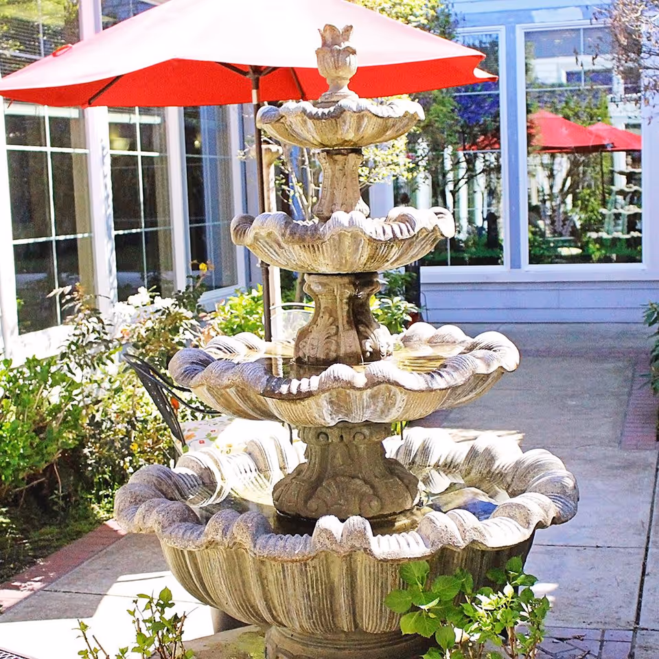 A multi-tiered stone water fountain in an outdoor courtyard area with red umbrellas, greenery, and a building with large windows in the background.