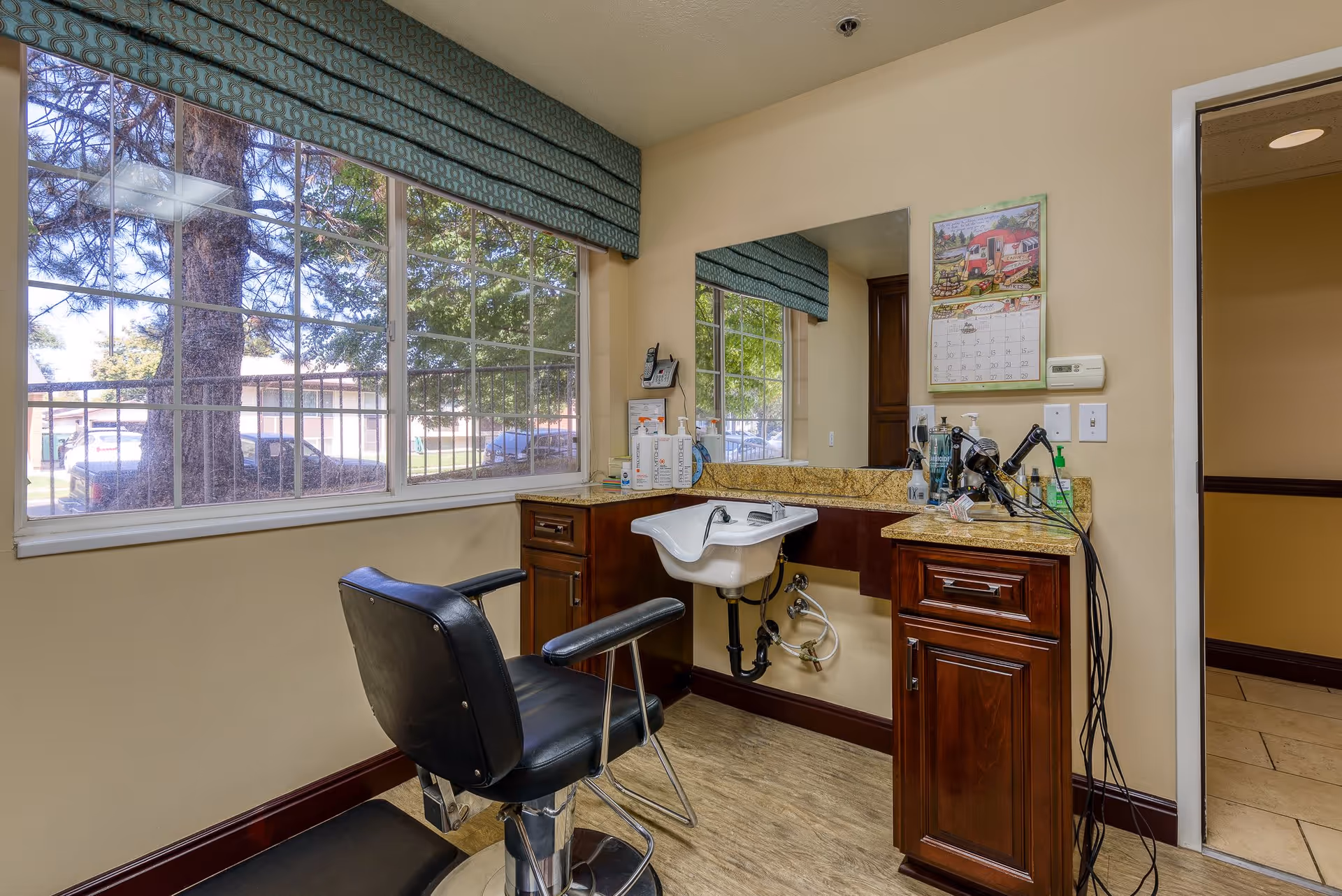 Interior view of a hair salon area in a senior living facility with a black salon chair in front of a small white sink. The room has large windows with a view of trees and parked cars outside. There are wooden cabinets with granite countertops, various hair care products, and a calendar on the wall.