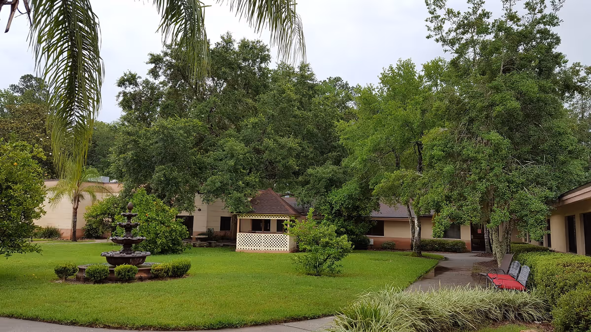 A lush green courtyard with a multi-tiered water fountain surrounded by small bushes. There are tall trees and a small gazebo with lattice walls in the background. The courtyard is bordered by a beige building with windows and a pathway with benches along the side.