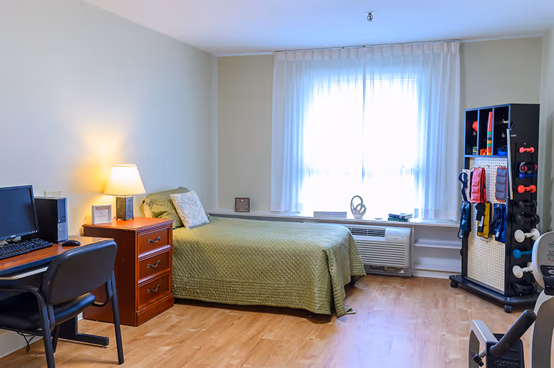 A senior living bedroom with a single bed covered in a green quilt, a wooden nightstand with a lamp, a desk with a computer and chair, a window with sheer white curtains, and a rack holding various exercise equipment including dumbbells and resistance bands.