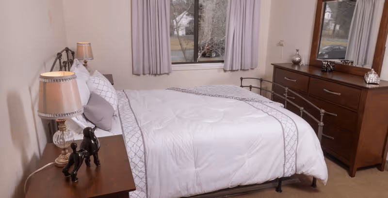 A neatly made bed with white and gray bedding in a senior living bedroom. The room features a wooden nightstand with a lamp and decorative black dog figurines, a window with gray curtains, and a wooden dresser with a mirror and decorative items on top.