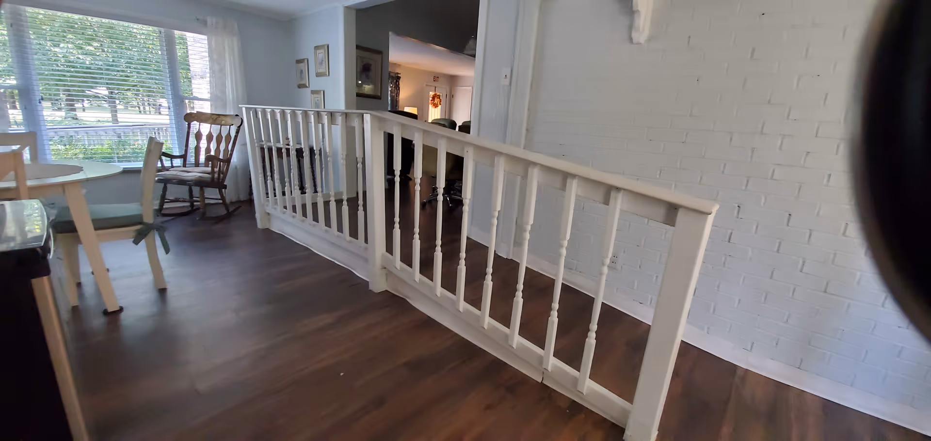 Interior living area with a white spindle railing, dark wood floors, a rocking chair and a small table by a large window.
