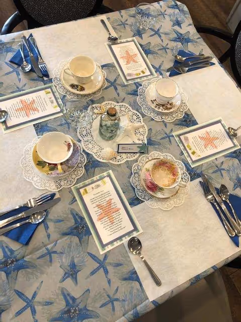 A dining table set for four with a blue and white starfish-themed tablecloth. Each place setting includes a teacup on a lace doily, a menu card, and silverware wrapped in blue napkins. In the center of the table is a small decorative bottle and a name card.