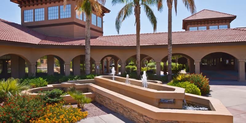 Entrance of a senior living facility with a tiered stone fountain, palm trees, arched colonnade and red tile roof.