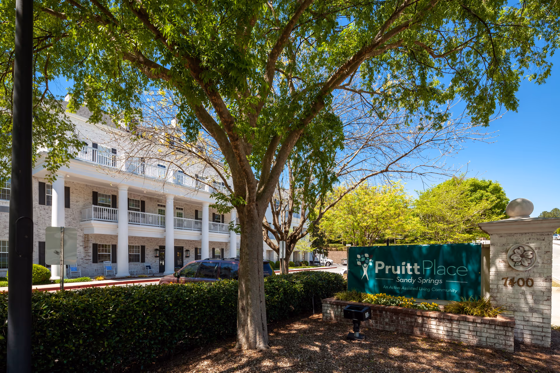 Exterior view of PruittPlace - Sandy Springs, an assisted living community, showing a large white building with columns and balconies. There is a green sign in front with the facility name and address number 7400. Trees and shrubs surround the area under a clear blue sky.