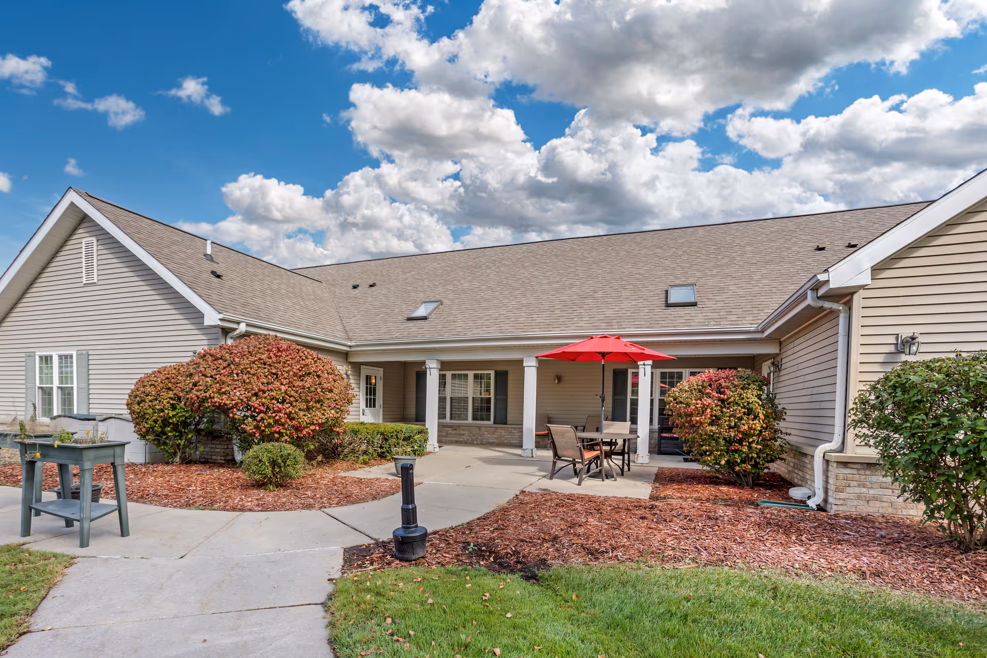 Single-story senior living building with a covered patio, outdoor table and red umbrella, shrubs, and a walkway under a partly cloudy sky.
