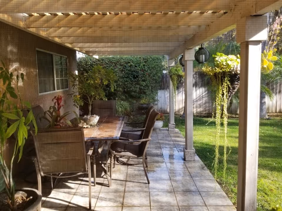 Outdoor patio area with a wooden pergola overhead, a tiled floor, a wooden table surrounded by chairs, potted plants, hanging planters, and a green lawn with bushes and trees in the background.