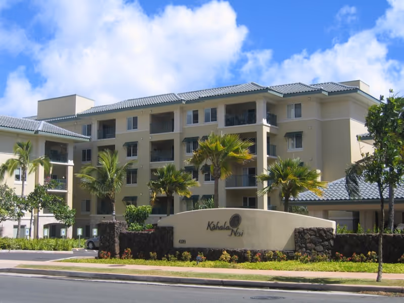 Exterior view of a multi-story senior living facility named Kahala Nui with beige walls, blue-gray tiled roof, palm trees, and a landscaped entrance sign.