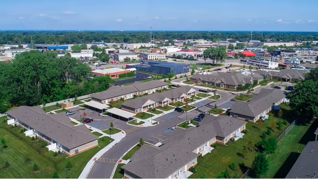 Aerial view of a senior living facility with multiple single-story buildings arranged along curved roads, surrounded by green lawns and trees, with a commercial area visible in the background under a clear blue sky.