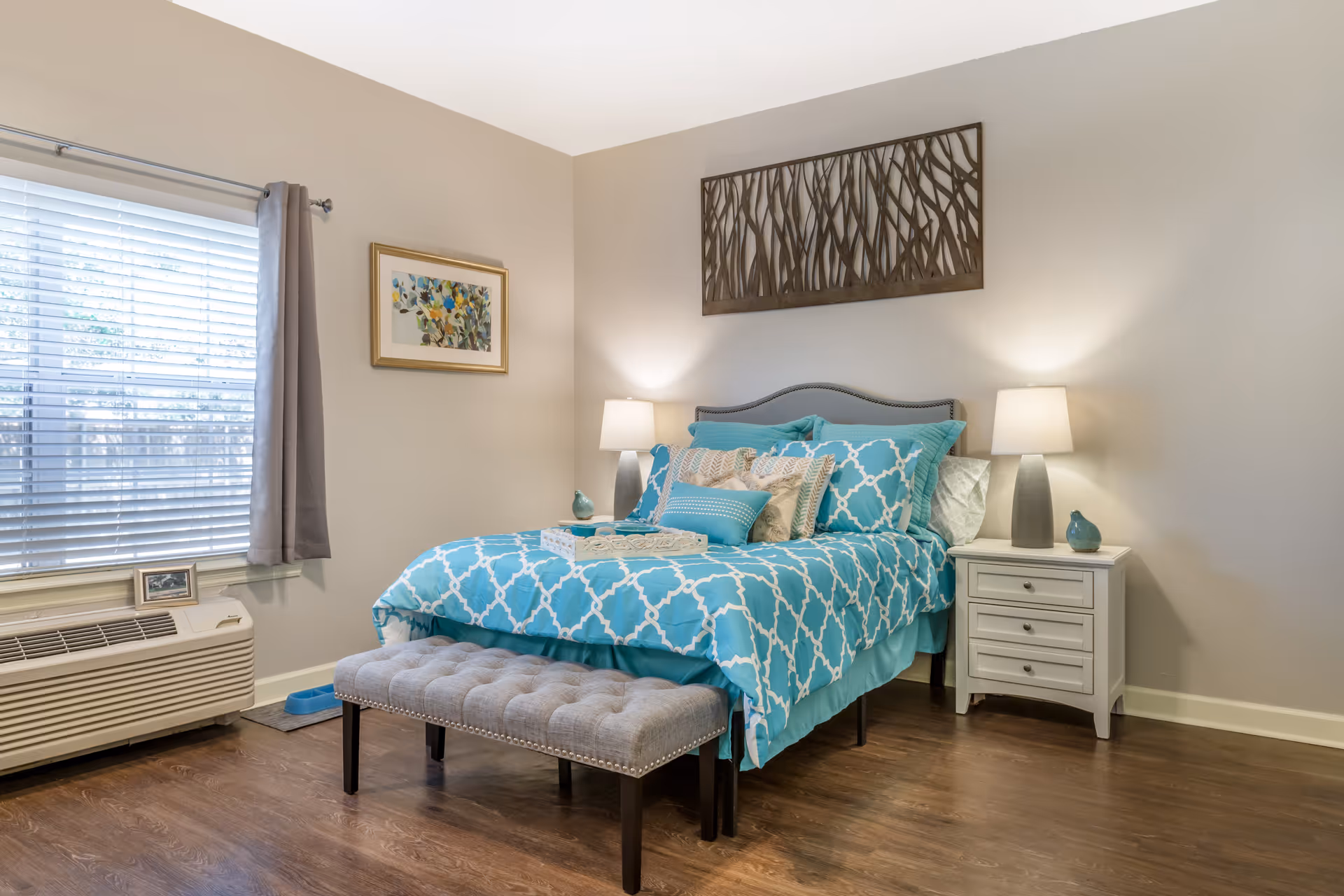 A bedroom with a bed dressed in turquoise and white patterned bedding, multiple pillows, and a tufted bench at the foot of the bed. There are two bedside tables with lamps and decorative items. A window with blinds and gray curtains is on the left wall, and two pieces of wall art hang above and beside the bed. The room has wooden flooring and neutral-colored walls.