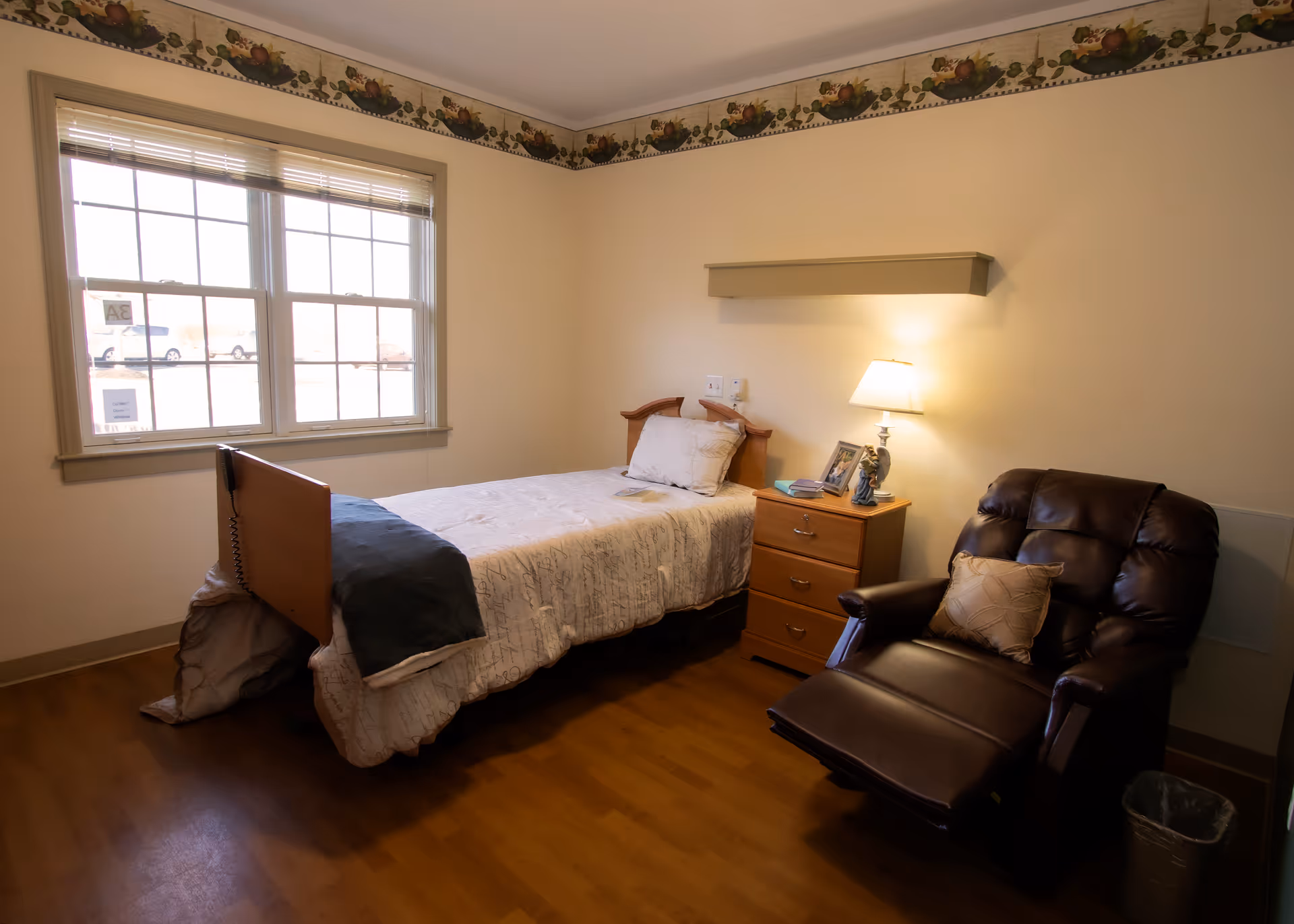 A small bedroom with a single bed covered in a light-colored bedspread, a wooden nightstand with a lamp, picture frames, and a figurine, and a dark brown recliner chair with a pillow. There is a large window with blinds letting in natural light, and a decorative border with fruit motifs runs along the top of the walls.