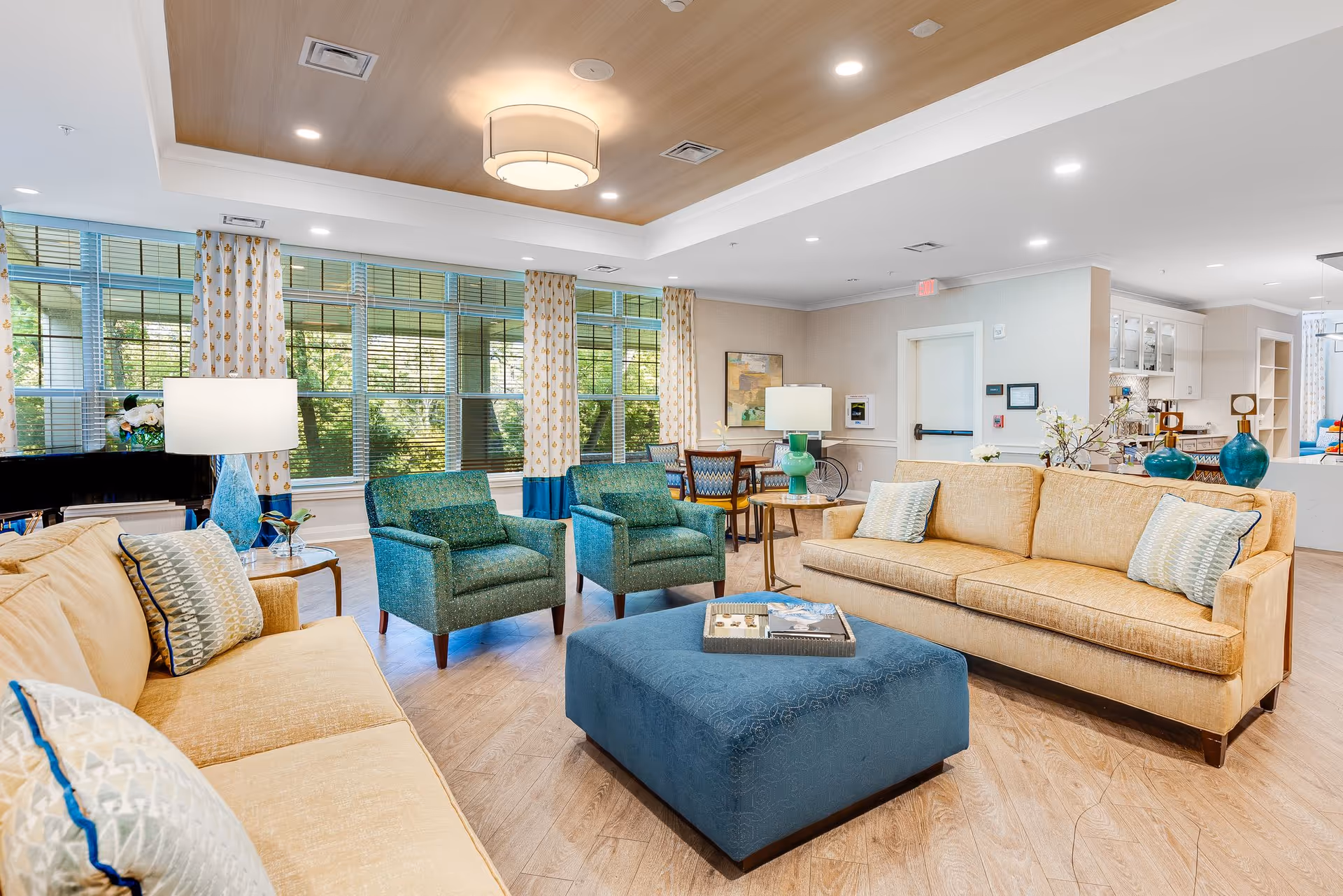 Bright communal living room with sofas, armchairs, a large ottoman, lamps, and large windows overlooking greenery.
