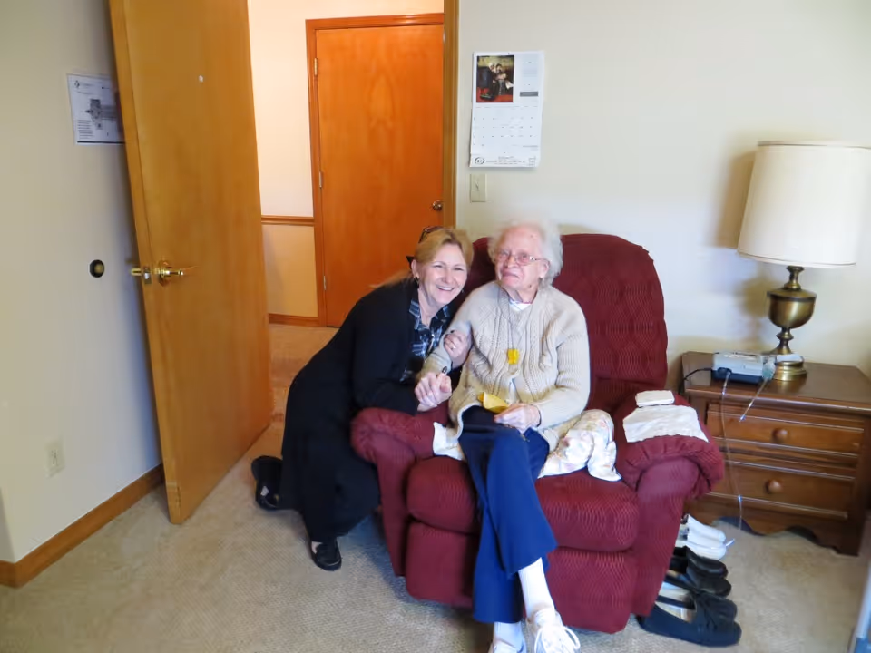 An elderly woman sitting in a red armchair inside a room, with a younger woman kneeling beside her, both smiling. The room has beige walls, a wooden door, a nightstand with a lamp, and a calendar on the wall.