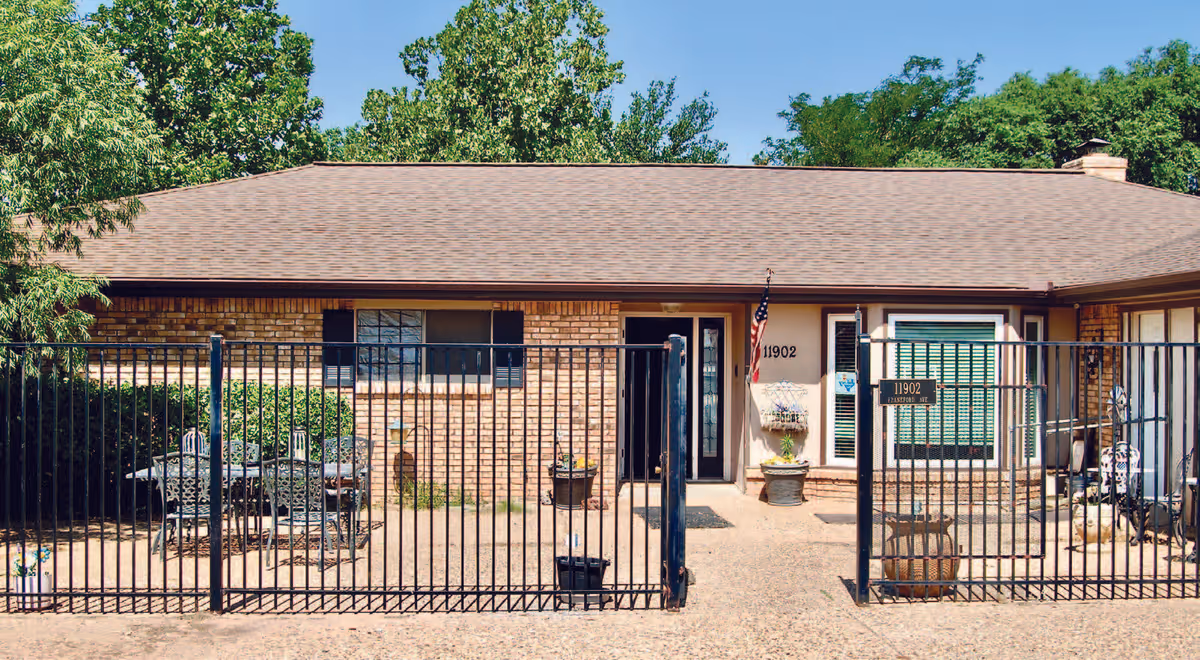 Single-story brick building with a brown shingled roof, black metal fence in front, and a small patio area with outdoor furniture. The building has windows with shutters, a door with the number 11902, and an American flag mounted near the entrance. Trees and greenery are visible in the background under a clear blue sky.