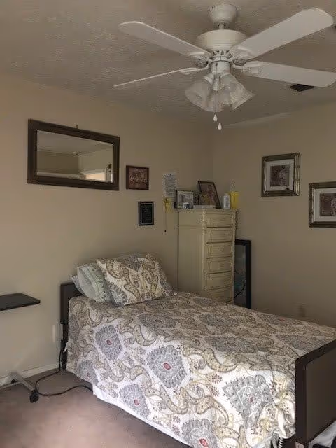 A small bedroom with a single bed covered in a patterned bedspread and matching pillow. Above the bed is a large mirror on the wall. To the right of the bed is a tall cream-colored dresser with various items on top, including framed photos and a yellow bottle. The walls are beige, and there is a ceiling fan with lights above the bed. Two framed pictures hang on the wall to the right of the dresser.