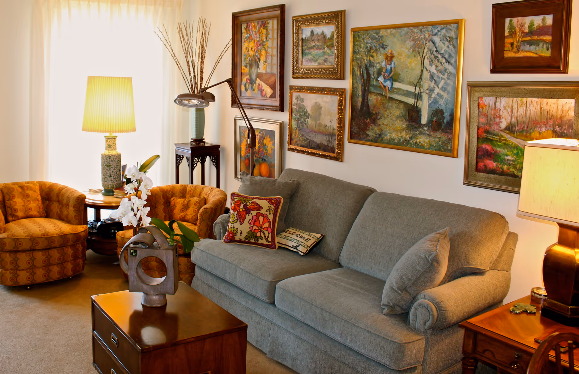A cozy living room with a gray sofa adorned with decorative pillows, two orange patterned armchairs, a wooden coffee table with a modern sculpture and white orchids, two table lamps providing warm light, and a wall decorated with multiple framed paintings depicting nature and floral scenes.